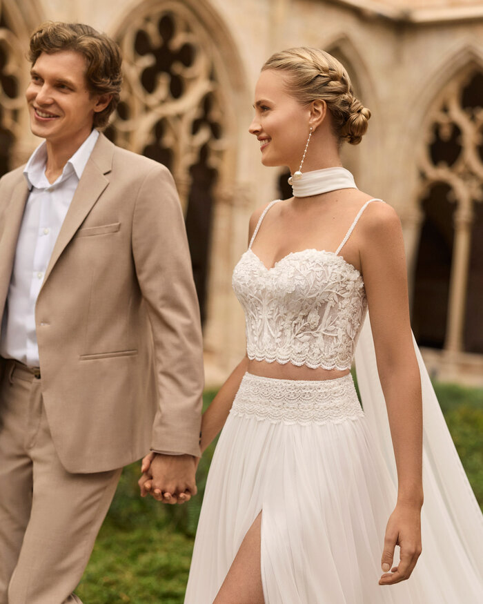 Nahaufnahme eines Brautpaares, die Hand in Hand vor einer Kathedrale entlang spazieren, sie trägt das zweiteilige Brautkleid Livy 