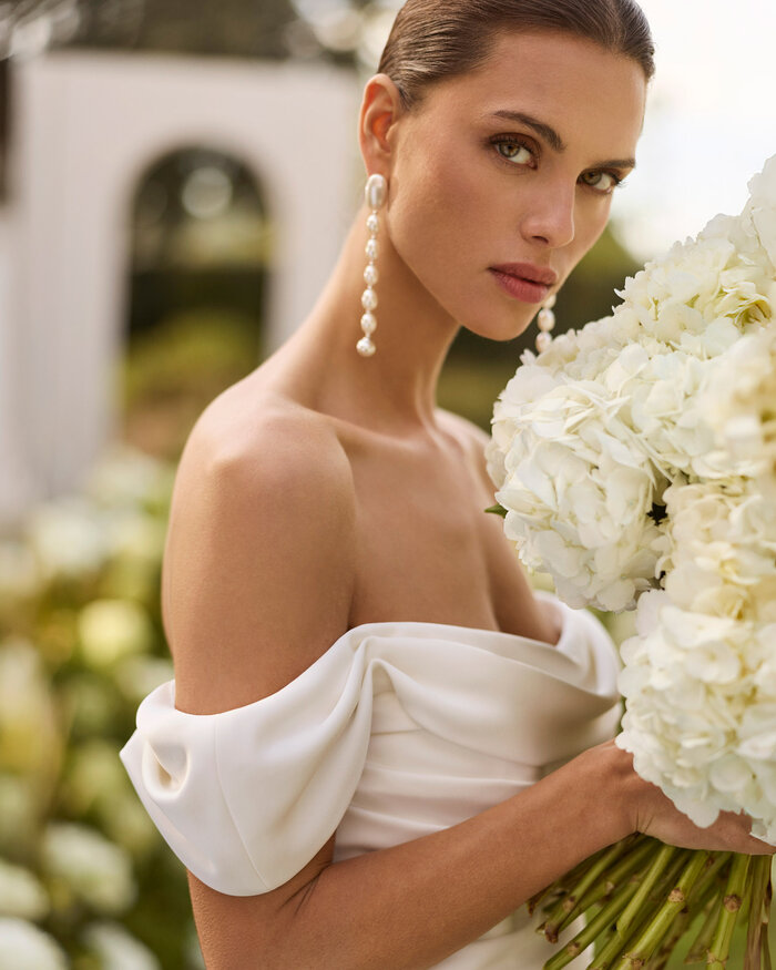 Das Brautkleid Nahe von Rosa Clara Nahaufnahme vom Brautkleid Nahe von Rosa Clará in Ivory mit schulterfreiem Drapé-Ausschnitt, langen transparenten Spitzenärmeln, figurbetonter Satin Silhouette und eleganter Schleppe, präsentiert in einem blühenden Garten.