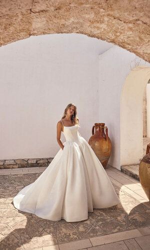 Brautkleid Lyrelle von Modeca Ganzkörperaufnahme einer Braut draußen vor einem Gewölbe, sie trägt das Kleid Lyrelle von Modeca und steckt ihre Hände in die Taschen
