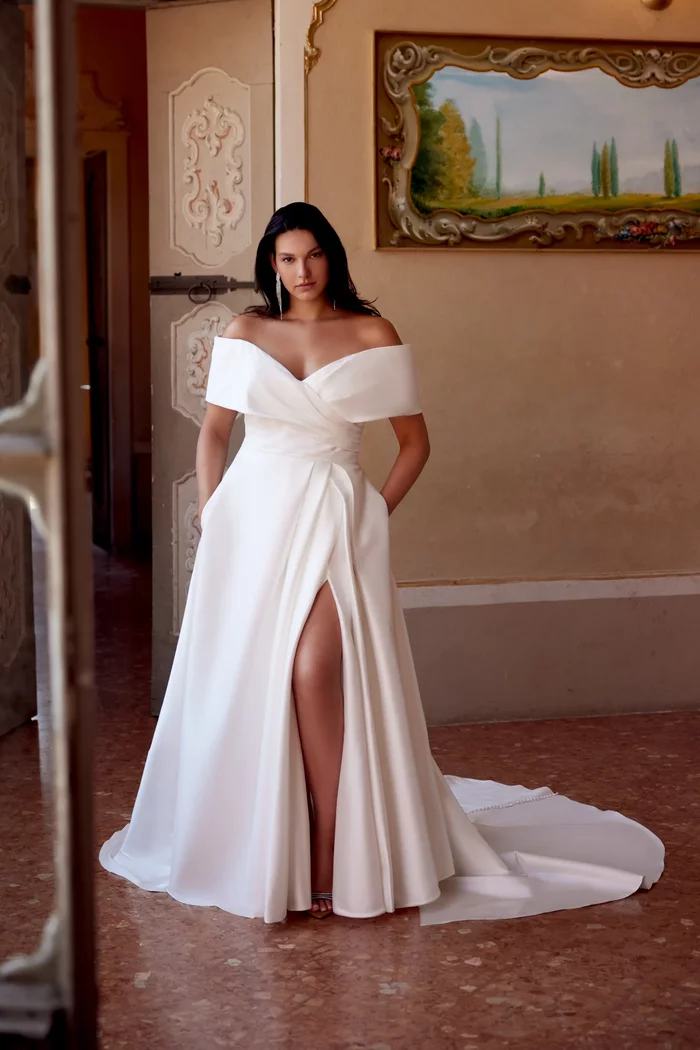 Curvy Brautkleid Harper von Justin Alexander in Ivory mit schulterfreiem Carmen-Ausschnitt, elegantem Wickeldesign, Beinschlitz und fließendem A-Linien-Rock mit schulterfreier Manschette – modernes Plus-Size Hochzeitskleid für stilvolle Bräute.