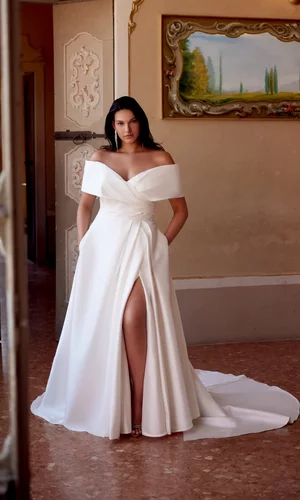 Curvy Brautkleid Harper von Justin Alexander in Ivory mit schulterfreiem Carmen-Ausschnitt, elegantem Wickeldesign, Beinschlitz und fließendem A-Linien-Rock mit schulterfreier Manschette – modernes Plus-Size Hochzeitskleid für stilvolle Bräute.