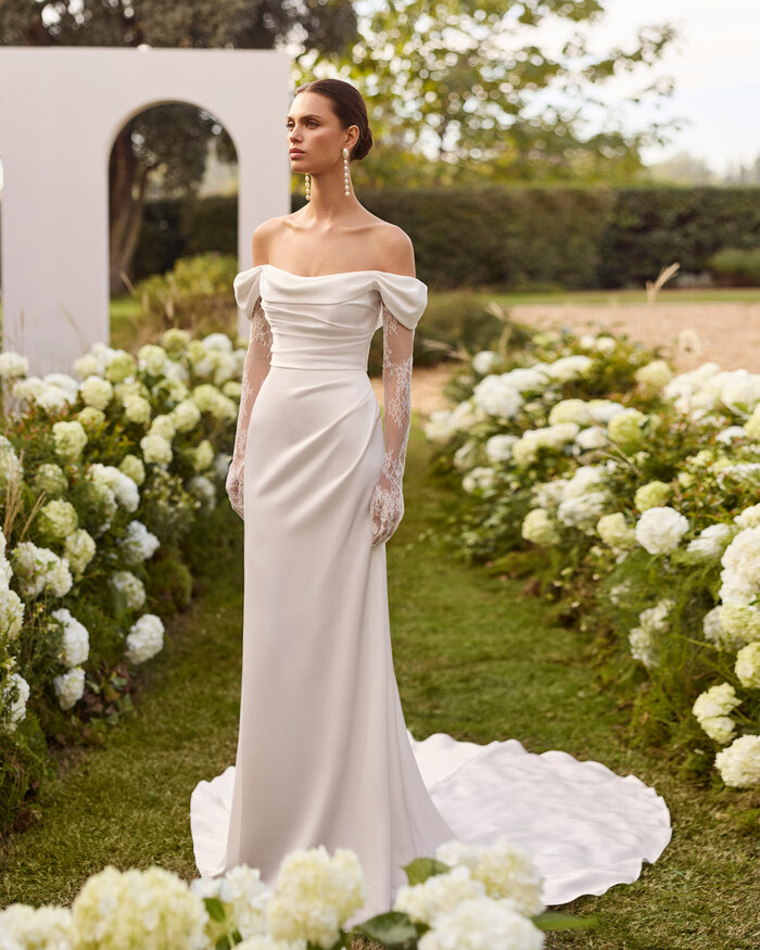 Das Brautkleid Nahe von Rosa Clara Brautkleid Nahe von Rosa Clará in Ivory mit schulterfreiem Drapé-Ausschnitt, langen transparenten Spitzenärmeln, figurbetonter Satin Silhouette und eleganter Schleppe, präsentiert in einem blühenden Garten.