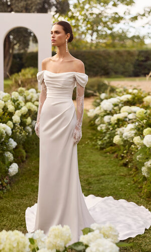 Das Brautkleid Nahe von Rosa Clara Brautkleid Nahe von Rosa Clará in Ivory mit schulterfreiem Drapé-Ausschnitt, langen transparenten Spitzenärmeln, figurbetonter Satin Silhouette und eleganter Schleppe, präsentiert in einem blühenden Garten.