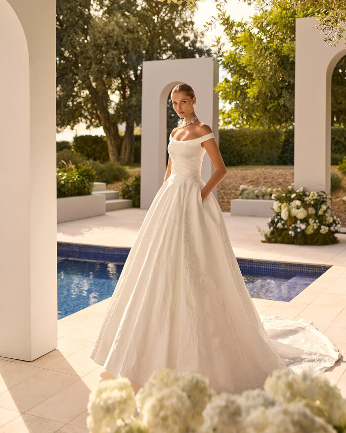 Das Brautkleid Nupcia von Rosa Clara Brautkleid Nupcia von Rosa Clará mit elegantem Off-Shoulder-Ausschnitt, weitem Rock und dezenter Blütenstickerei in einem Brokat Stoff