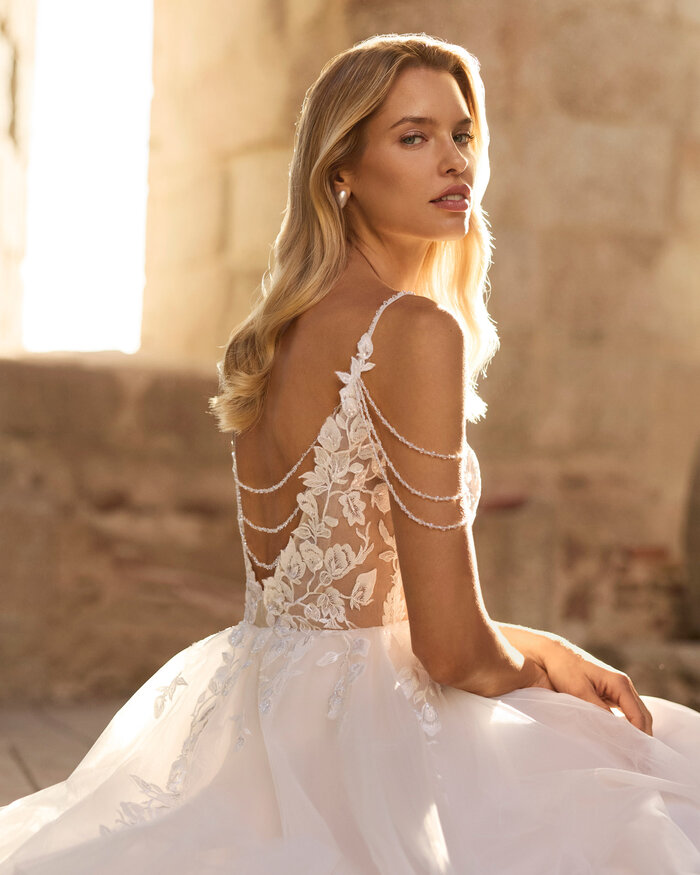 Brautkleid von Pinara von Adriana Alier Nahaufnahme einer Braut vor antiken Steinsäulen, sie trägt das Kleid Pinara von Adriana Alier, die Sonne scheint von hinten, ihr seitlicher Oberkörper wird gezeigt und sie schaut über die Schulter