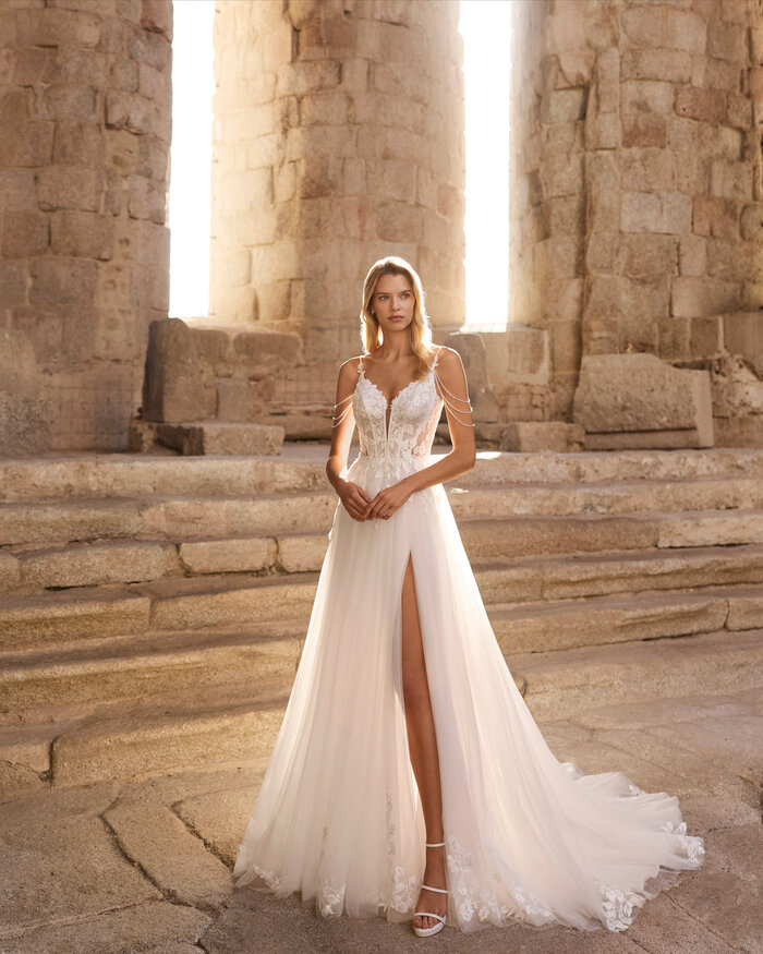 Brautkleid Pinara von Adriana Alier Ganzkörperaufnahme einer Braut vor antiken Steinsäulen, sie trägt das Kleid Pinara von Adriana Alier, die Sonne scheint von hinten, Braut zeigt den Schlitz im Kleid