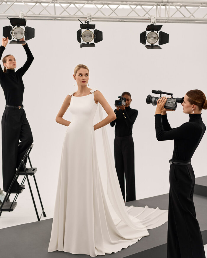 Ganzkörperaufnahme einer Braut in einem Studio, sie trägt das Kleid Scarlet von Adriana Alier, 3 in schwarz gekleidete Fotografinnen richten Kameras und Scheinwerfer auf die Braut 