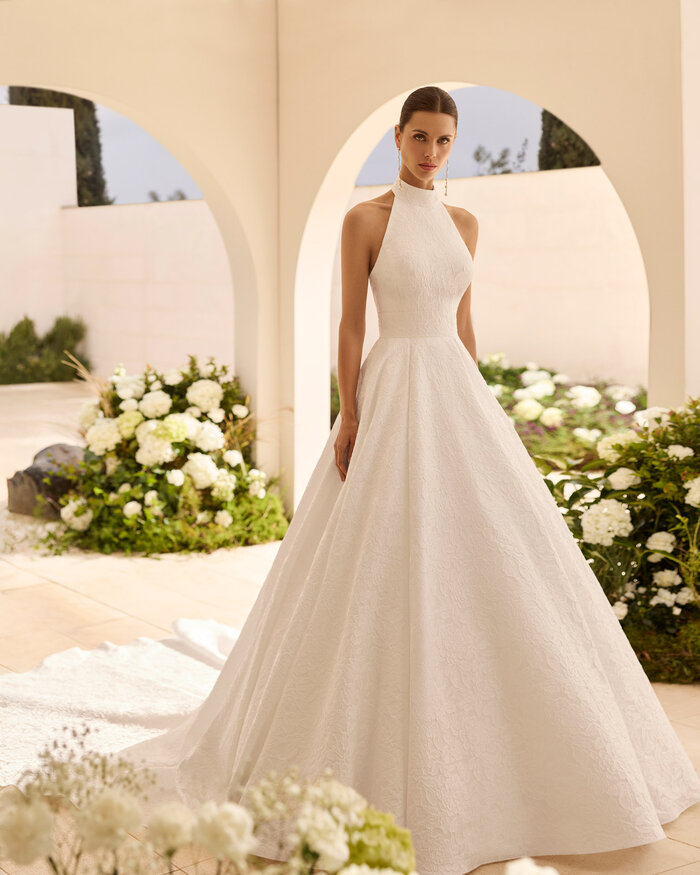 Das Brautkleid Nut von Rosa Clara Brautkleid Nut von Rosa Clará mit hochgeschlossenem Neckholder-Ausschnitt, eleganter Taillenbetonung und weit ausgestelltem A-Linien-Rock aus strukturiertem Stoff