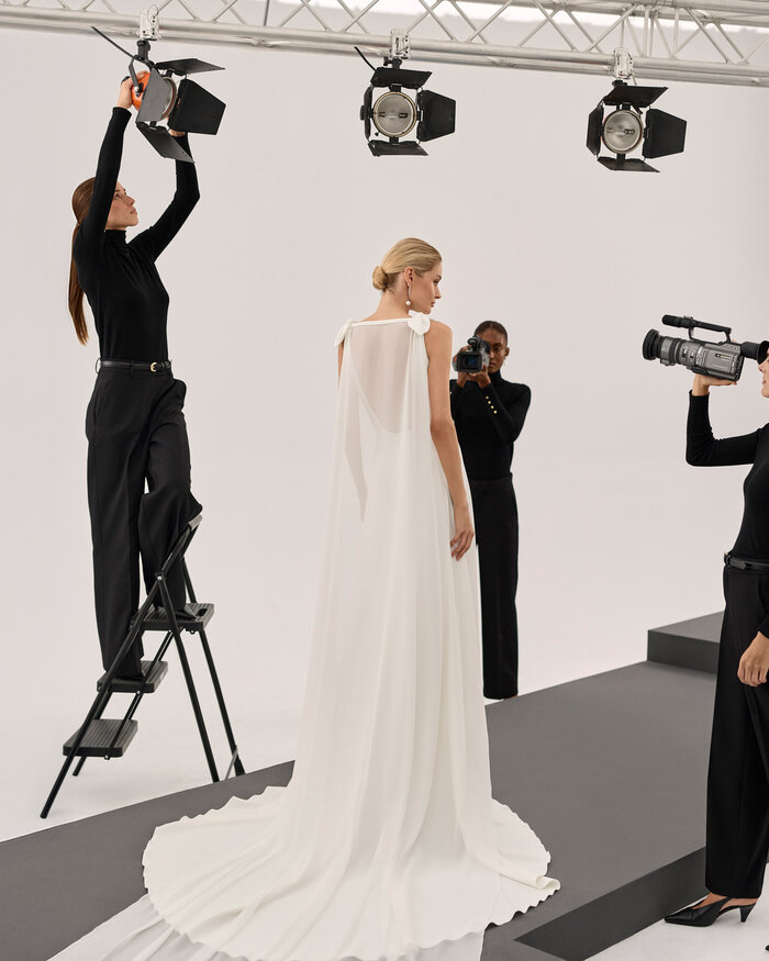 Ganzkörperaufnahme einer Braut von hinten in einem Studio, sie trägt das Kleid Scarlet von Adriana Alier und darüber einen transparenten Umhang, 3 in schwarz gekleidete Fotografinnen richten Kameras und Scheinwerfer auf die Braut 