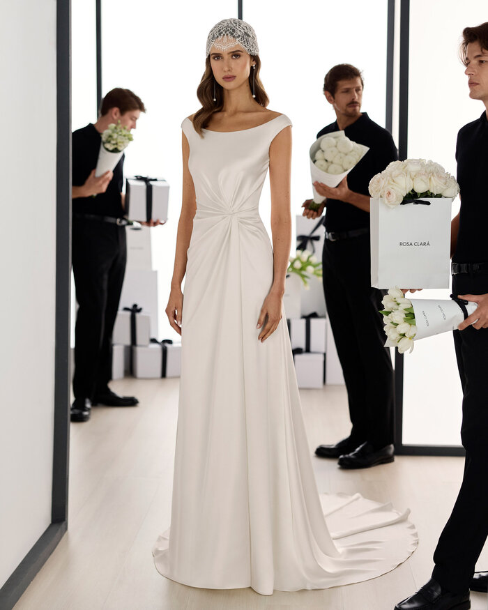 Ganzkörperaufnahme einer Braut im Studio, sie trägt das Brautkleid Solea von Adriana Alier, im Hintergrund befinden sich in schwarz gekleidete Männer die bei der Blumendeko helfen