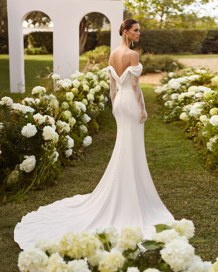 Das Brautkleid Nahe von Rosa Clara Rückenansicht vom Brautkleid Nahe von Rosa Clará in Ivory mit schulterfreiem Drapé-Ausschnitt, langen transparenten Spitzenärmeln, figurbetonter Satin Silhouette und eleganter Schleppe, präsentiert in einem blühenden Garten.