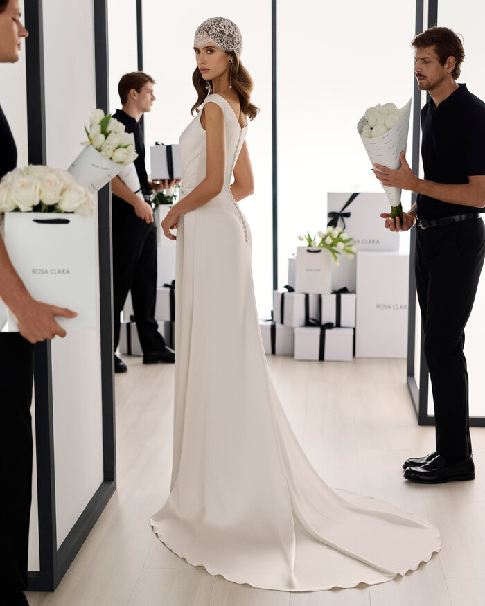 Ganzkörperaufnahme einer Braut von hinten im Studio, sie trägt das Brautkleid Solea von Adriana Alier, im Hintergrund befinden sich in schwarz gekleidete Männer die bei der Blumendeko helfen