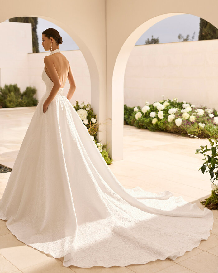 Das Brautkleid Nut von Rosa Clara Brautkleid Nut von Rosa Clará mit hochgeschlossenem Neckholder-Ausschnitt, eleganter Taillenbetonung und weit ausgestelltem A-Linien-Rock aus strukturiertem Stoff