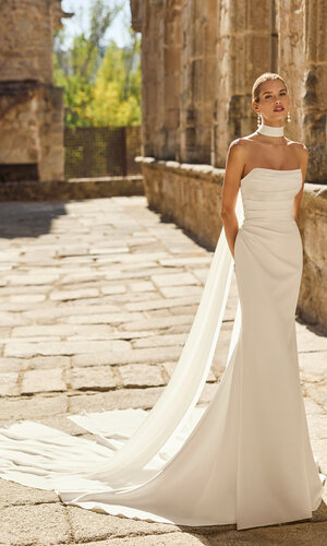 Brautkleid Paddy von Adriana Alier Braut trägt das moderne Brautkleid Paddy von Adriana Alier mit trägerlosem Ausschnitt, figurbetonter Fit-and-Flare-Silhouette und langer fließender Schleppe in historischer Kulisse.
