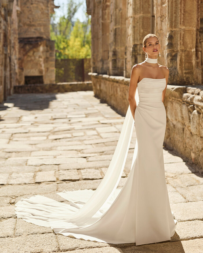 Brautkleid Paddy von Adriana Alier Braut trägt das moderne Brautkleid Paddy von Adriana Alier mit trägerlosem Ausschnitt, figurbetonter Fit-and-Flare-Silhouette und langer fließender Schleppe in historischer Kulisse.