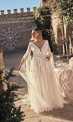 Das Brautkleid Nadia von Modeca Brautkleid Nadia von Modeca mit floraler Spitze, tiefem V-Ausschnitt und fließendem Tüllrock in einem romantischen Innenhof mit mediterranem Hochzeitsambiente.