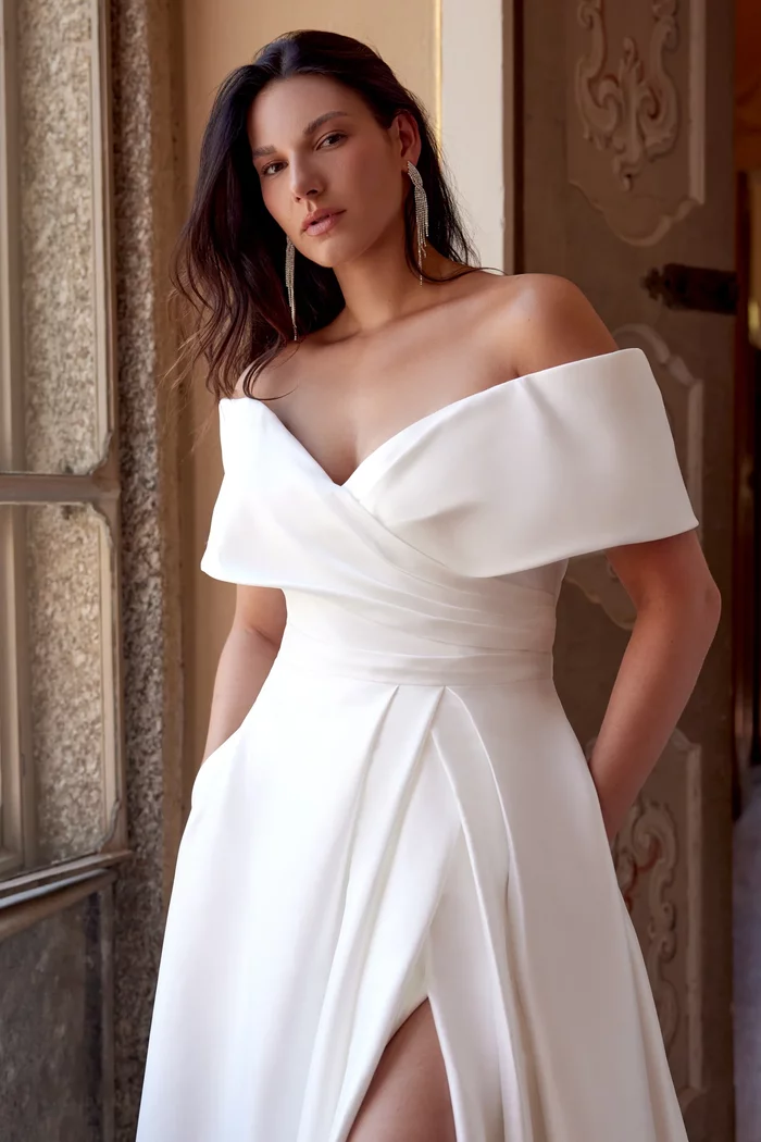 Curvy Brautkleid Harper von Justin Alexander in Ivory mit schulterfreiem Carmen-Ausschnitt, elegantem Wickeldesign, Beinschlitz und fließendem A-Linien-Rock mit schulterfreier Manschette – modernes Plus-Size Hochzeitskleid für stilvolle Bräute.